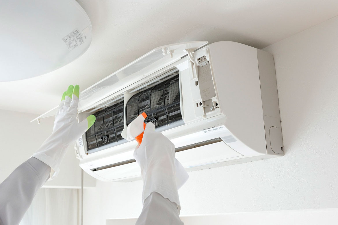 A technician wearing protective gloves sprays cleaner onto the filter of a wall-mounted air conditioner during AC repair and maintenance.