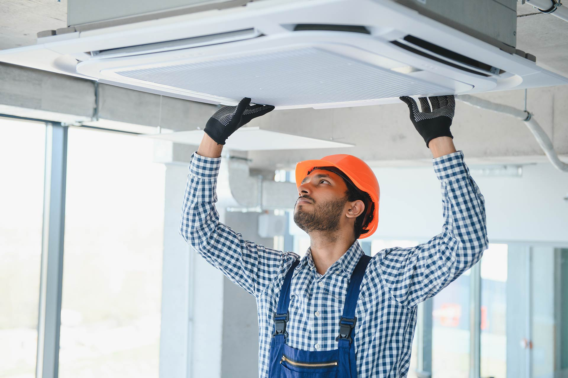 Technician wearing an orange safety helmet and gloves inspecting a ceiling-mounted central AC unit in a Dubai building.