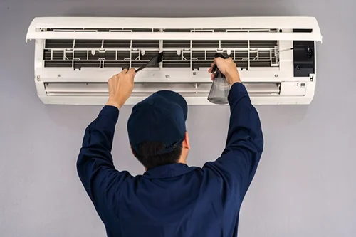 Technician in a blue uniform cleaning and repairing a wall-mounted air conditioner with a brush and spray bottle.